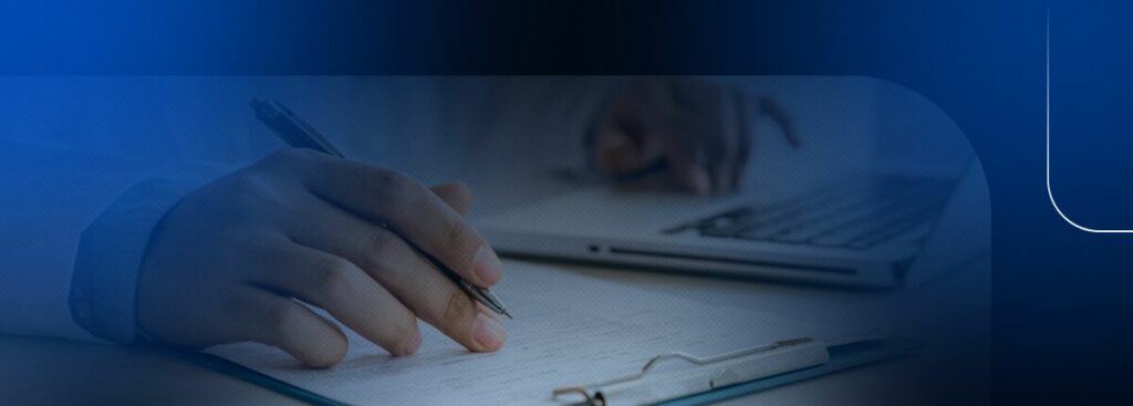 Pessoa escrevendo em um bloco de notas com laptop ao fundo